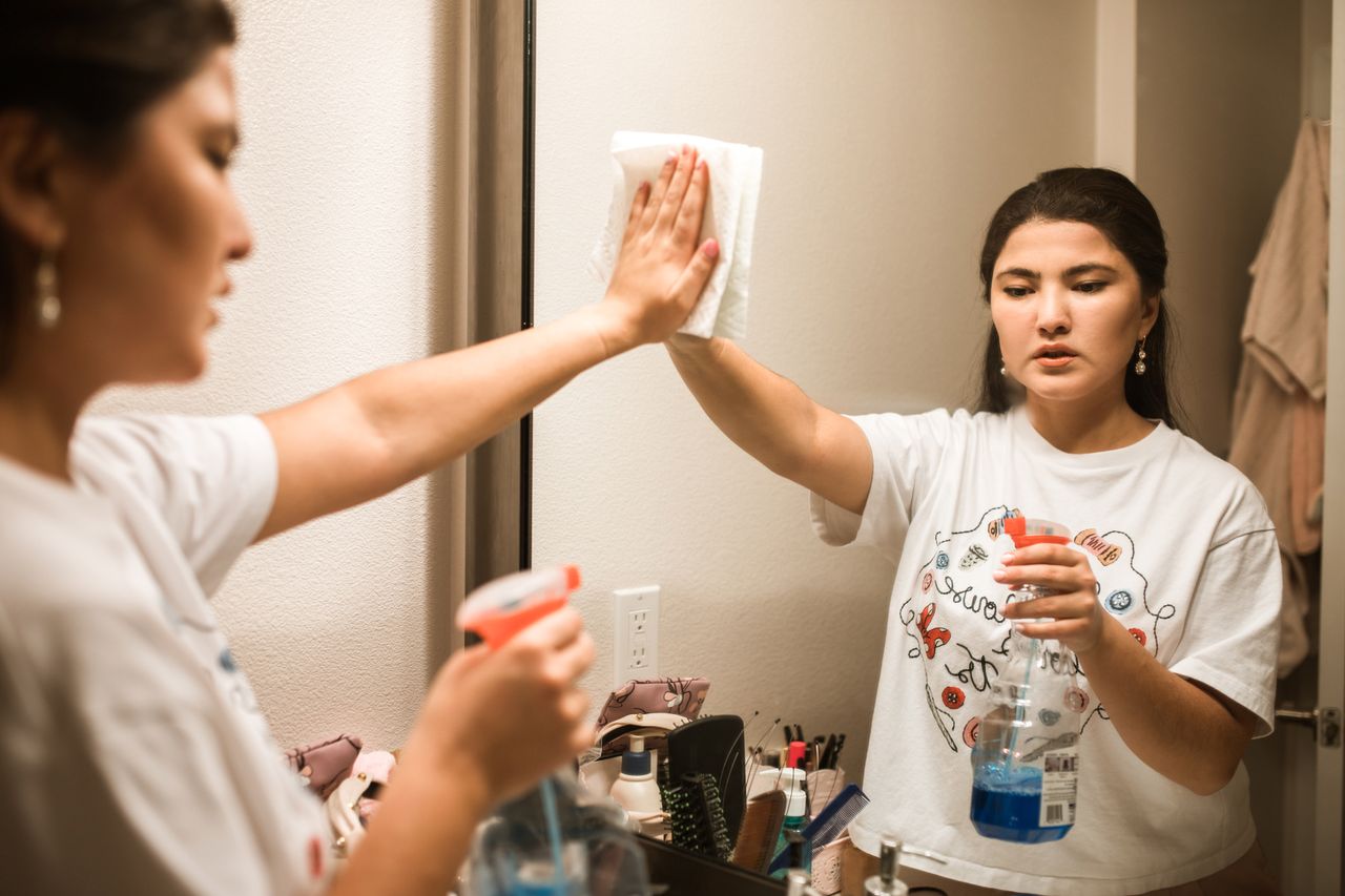 hero-img An adult woman cleaning a bathroom mirror with a spray bottle and cloth, focused on hygiene.
